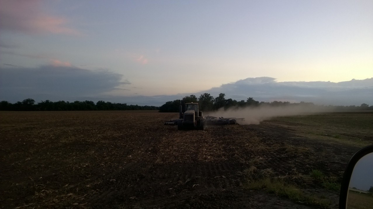 A tractor pulling a wide implement at dusk, dust trailing off behind it