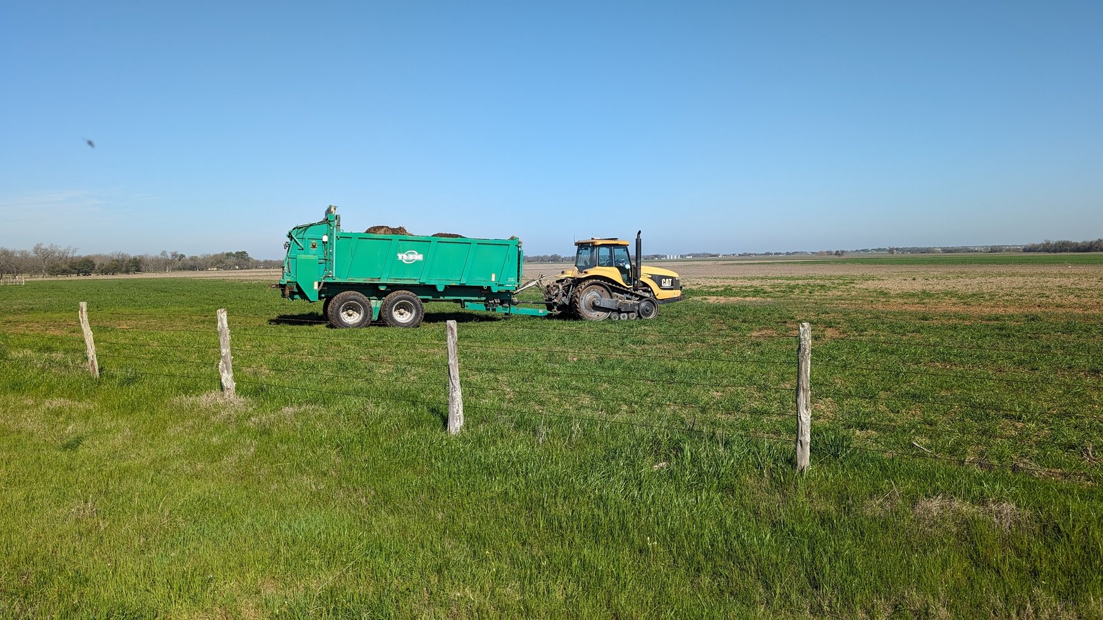 A grain cart behind a tracked tractor on green pasture, a fence line running past