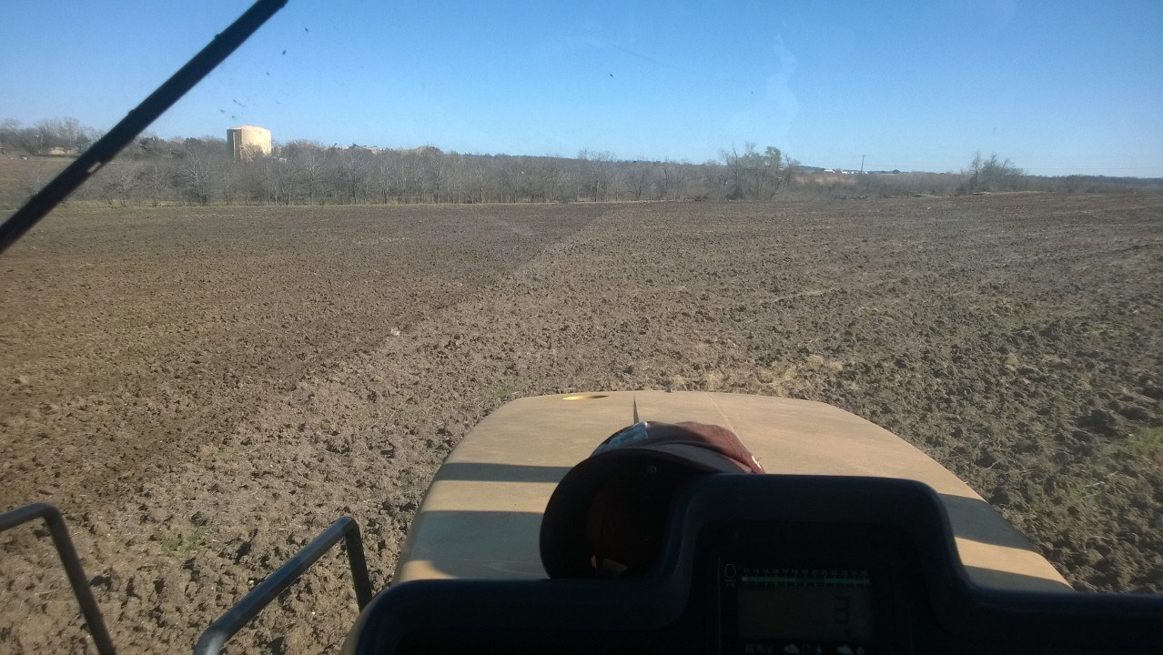 The view forward over the hood of a tractor across freshly worked ground, a guidance display on the console