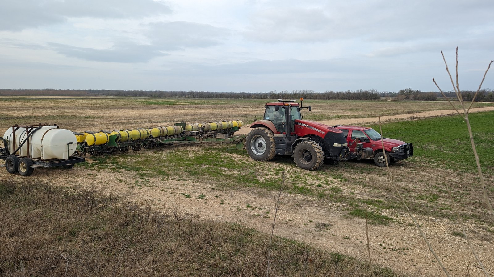 A Case IH tractor pulling a planter with its liquid tank, out on worked ground beside a tree line
