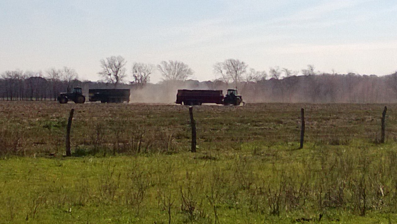 Two tractors pulling spreader boxes across a field, dust rising behind them, seen from beyond a fence